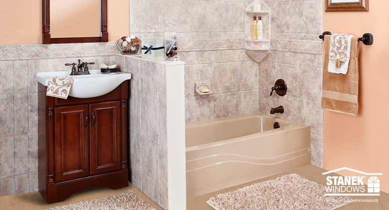 A newly remodeled bathroom with a sink in a wood cabinet, beige tub, peach walls, and white and gray tile on the some walls.